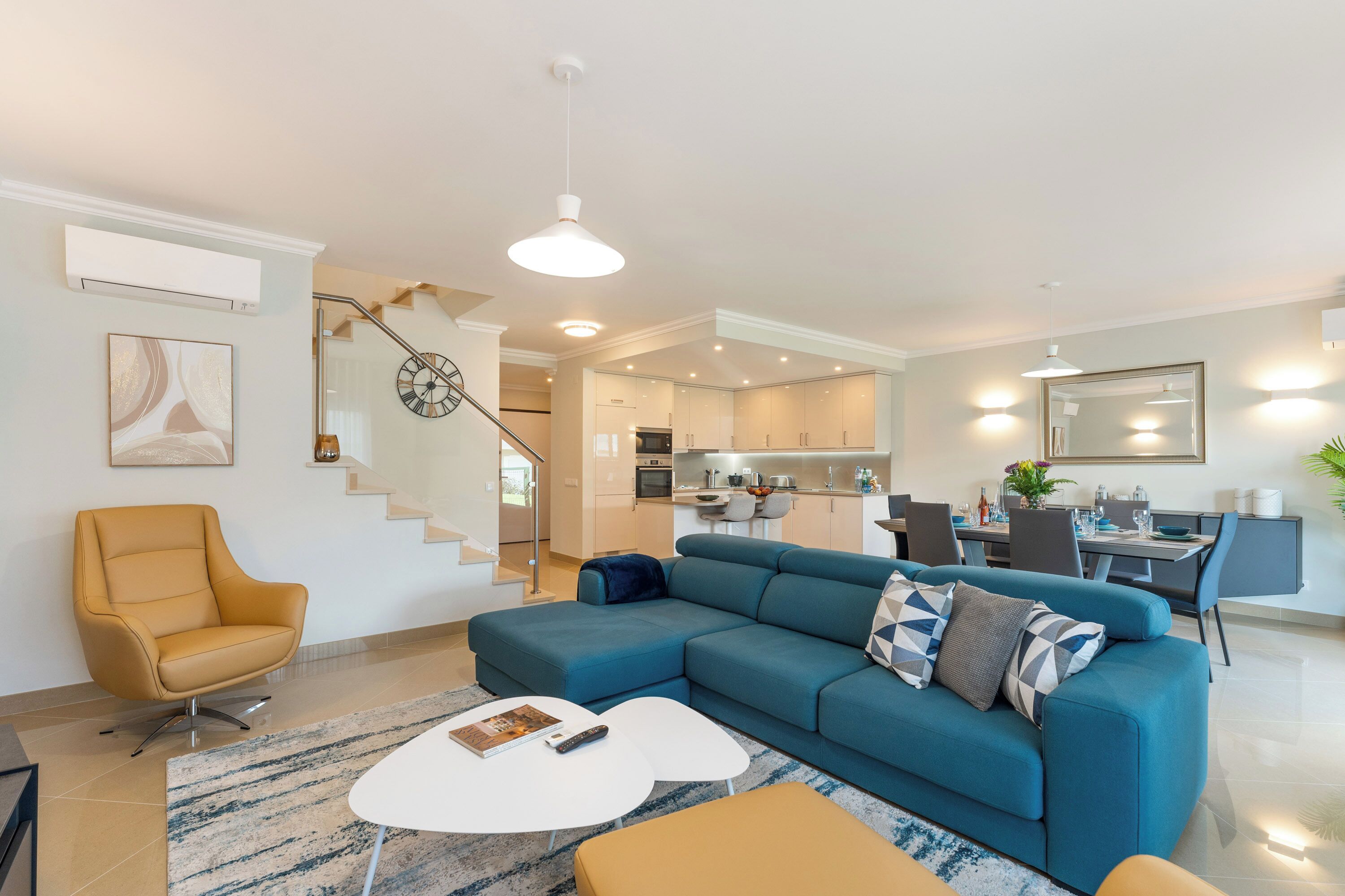 A modern living room with a blue sofa, a yellow armchair, and a white coffee table. A staircase leads to an open kitchen area.