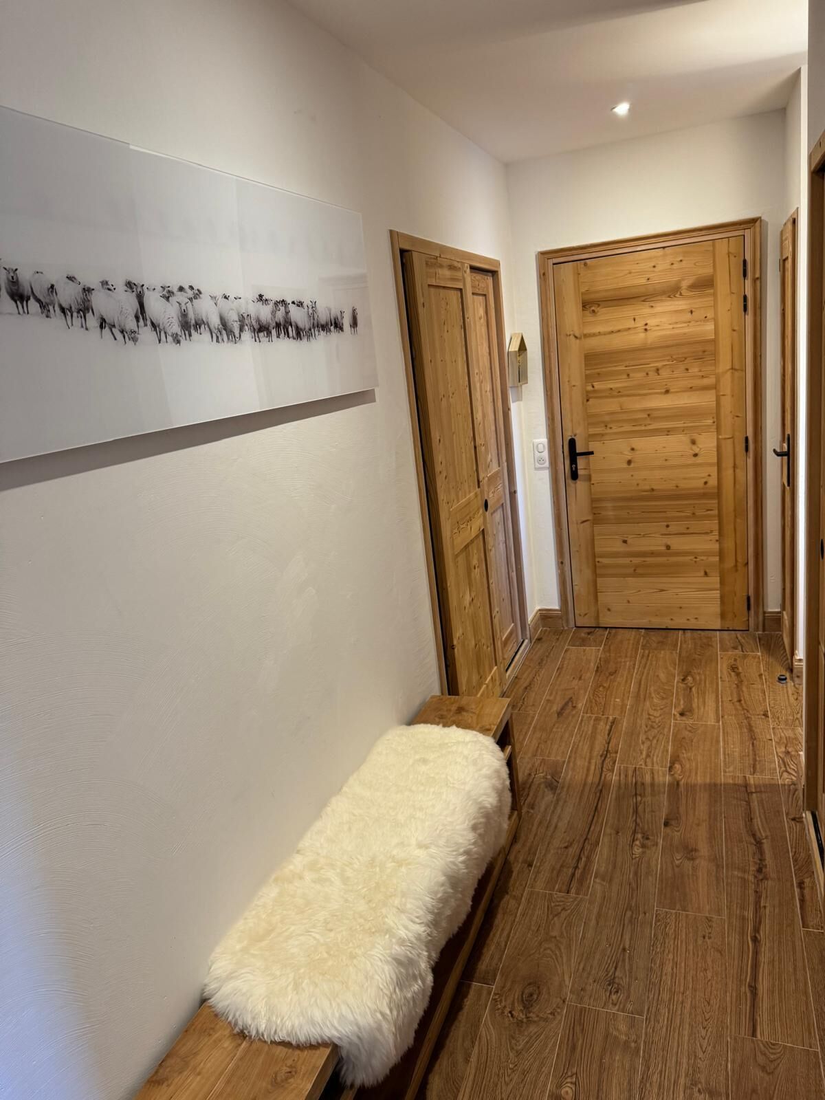 A hallway with wooden flooring, a bench with a fluffy white cover, and a wooden door.