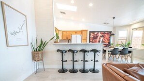 A modern kitchen with wooden cabinets, a white refrigerator, and a dining area with a table and chairs. There are four black bar stools in front of the kitchen.