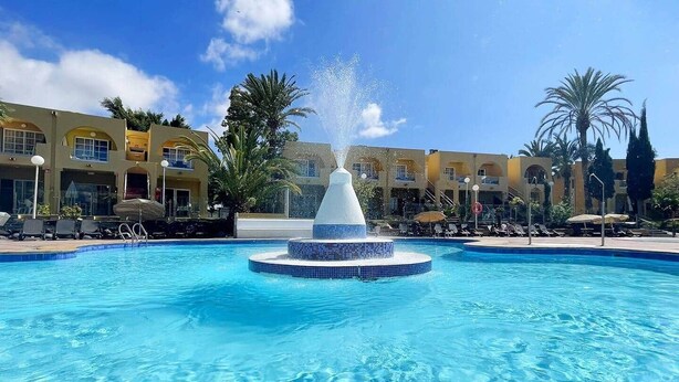 Una piscina con una fuente central, rodeada de sillas de descanso y palmeras.