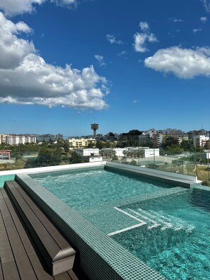 Una piscina al terrat amb aigua blava cristal·lina, envoltada per una terrassa de fusta i una vista panoràmica de la ciutat.