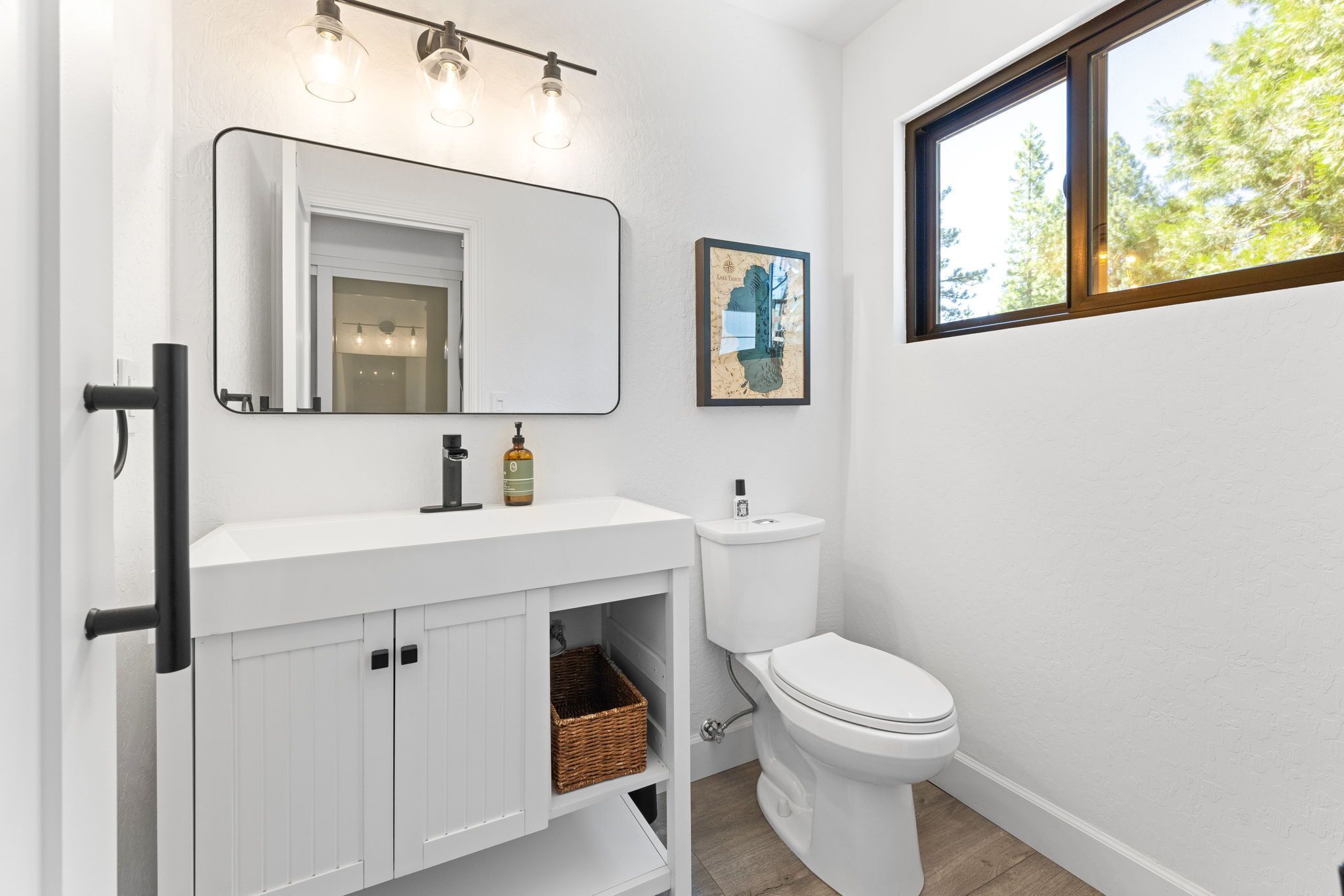 A bathroom with a white vanity, a large mirror, a toilet, and a window with a view of trees.