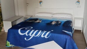 Quarto com cama coberta por um cobertor azul com a inscrição 'Capri', almofadas brancas e mesa de cabeceira com abajur.