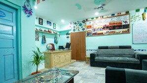 Un área de recepción con una puerta azul, un mostrador de recepción, un sofá gris, una mesa con cubierta de vidrio y objetos coloridos, y una pared con diversas decoraciones y una pizarra.