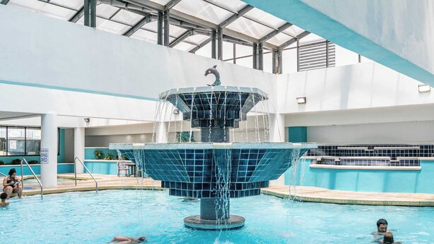 An indoor swimming pool with a central fountain, clear water, and a tiled deck surrounding the pool area. - Sky-High Oceanfront Stay Pools and DJs & More (Myrtle Beach)