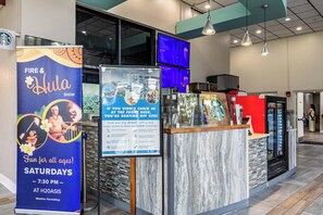 A hotel reception area with a large sign for the Fire & Hula Show on Saturdays, a digital menu board, and a Starbucks coffee machine. - Sky-High Oceanfront Stay Pools and DJs & More (Myrtle Beach)
