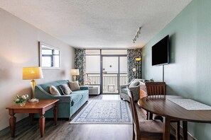 Uma sala de estar com sofá, mesa de jantar, TV e varanda com cortinas.