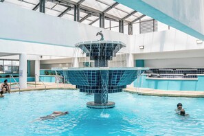 An indoor swimming pool with a central fountain, clear water, and a tiled deck surrounding the pool area. - Sky-High Oceanfront Stay Pools and DJs & More (Myrtle Beach)