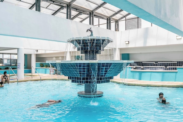An indoor swimming pool with a central fountain, clear water, and a tiled deck surrounding the pool area.