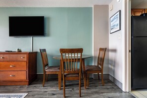 A room with a flat-screen TV mounted on the wall, a wooden dresser, two wooden chairs around a small round table, and a black refrigerator in the corner.