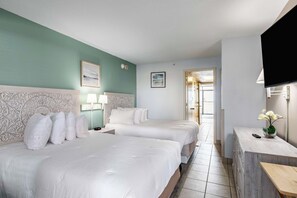 A hotel room with two beds, a TV, a desk with a vase of flowers, and a door leading to another room.