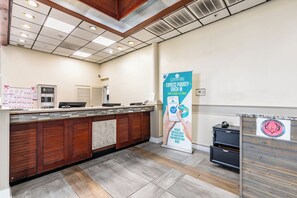 Uma área de recepção de hotel com um balcão de madeira, um quiosque digital de check-in, uma máquina de café e um banner promovendo check-in prioritário expresso.