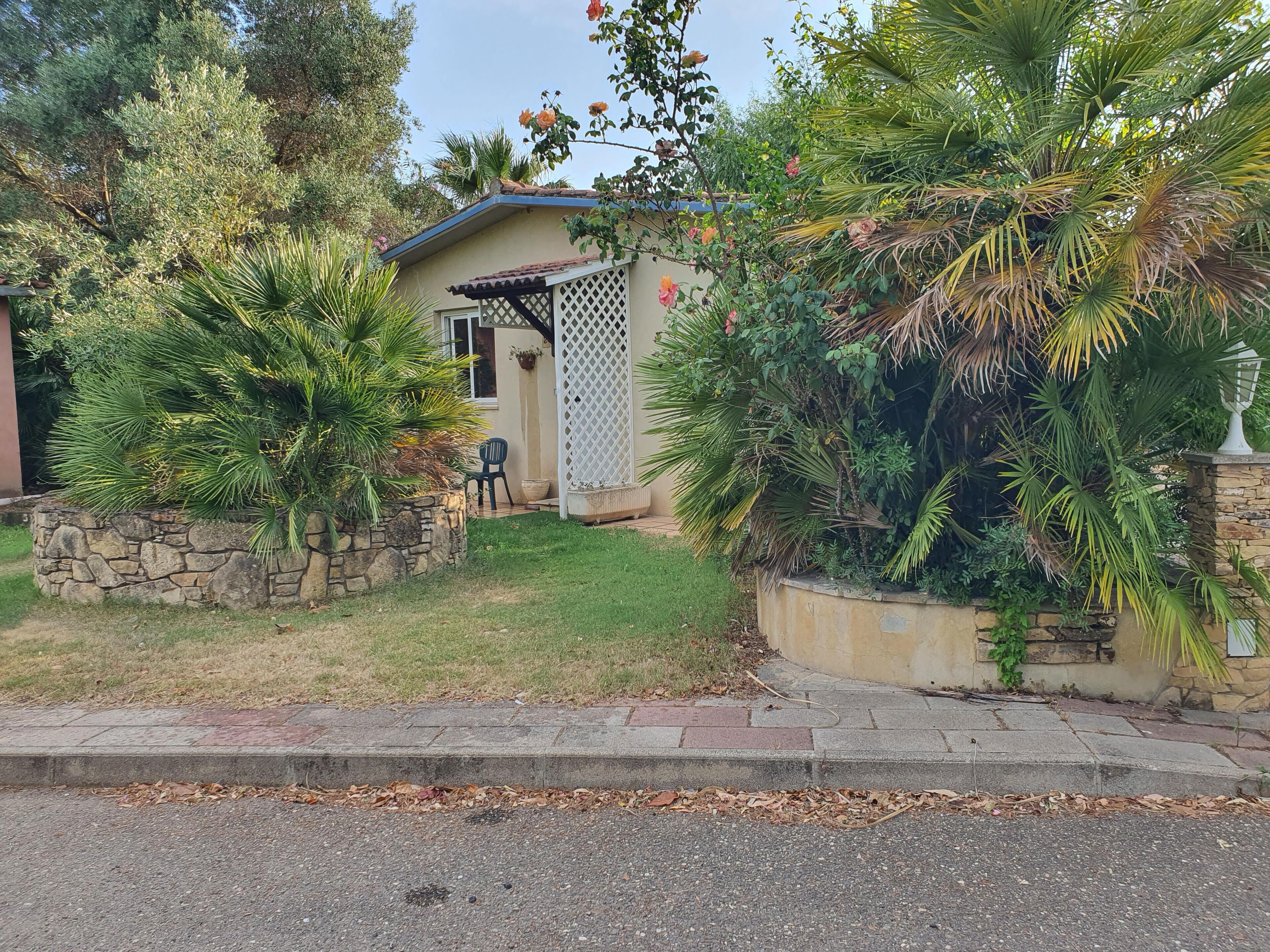 Una casa con un muro de piedra, un pequeño porche y diversas plantas, entre ellas palmeras y rosales.