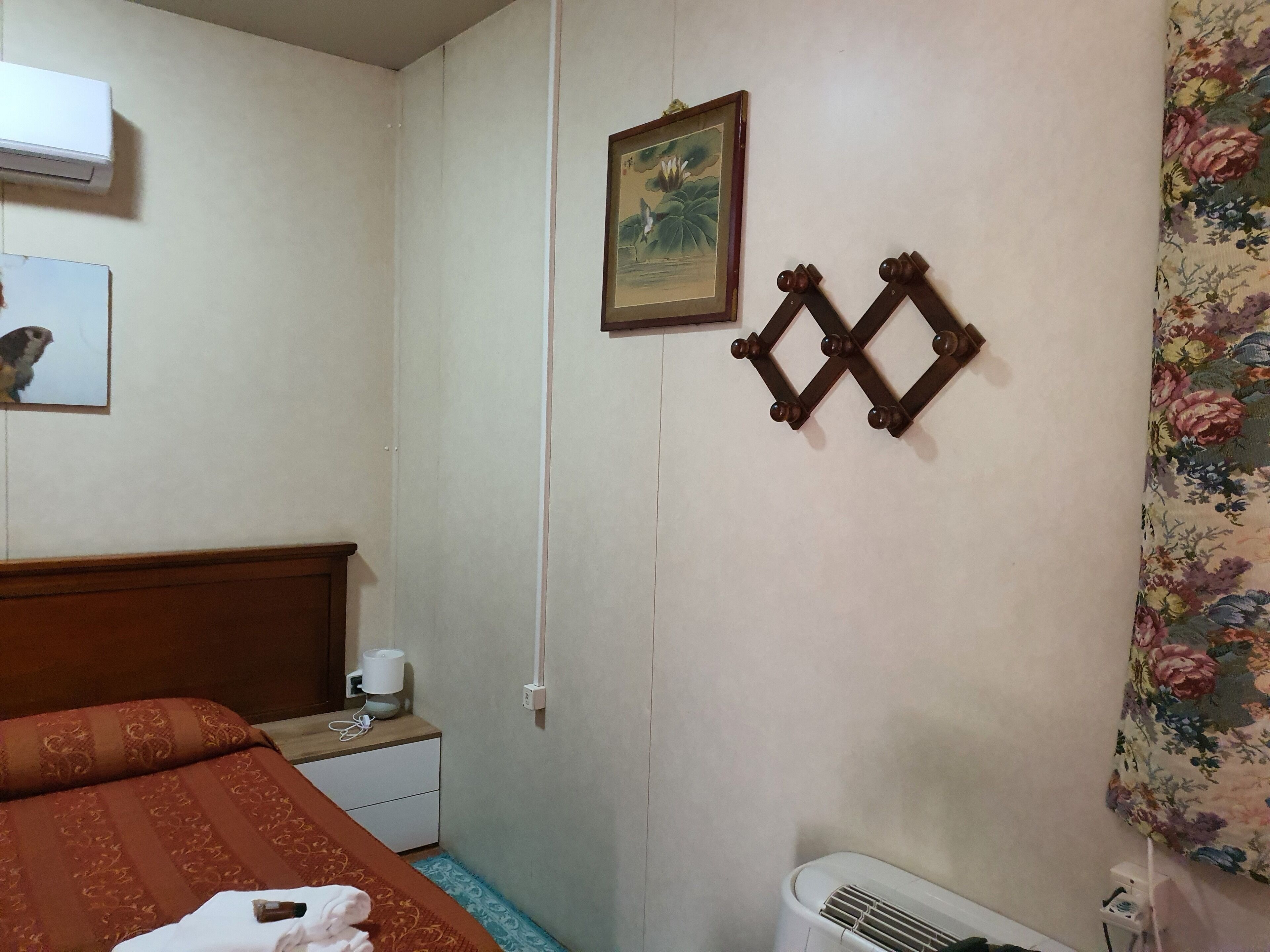 Una camera d'albergo con un letto, un comodino, una lampada, un portafoto, un elemento decorativo a parete e una tenda floreale.