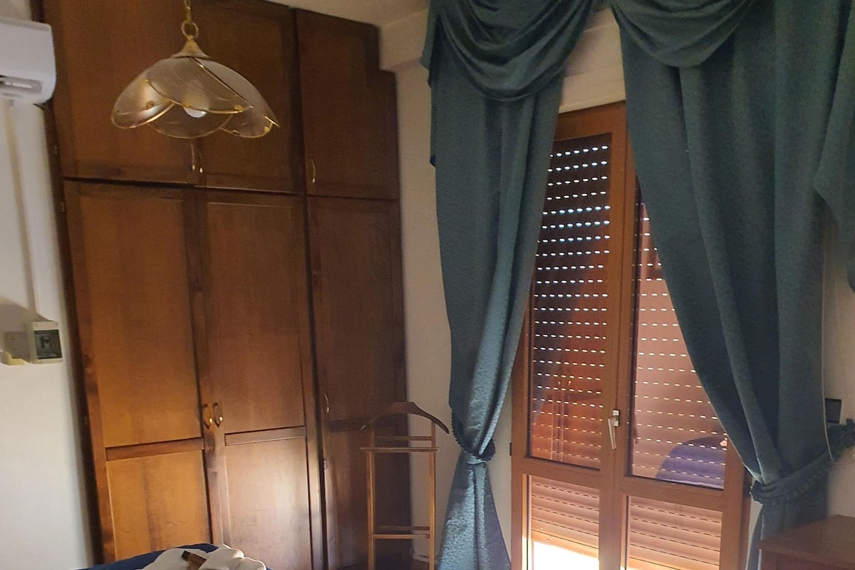 Ein Schlafzimmer mit einem Bett, einem Kleiderschrank, einer Hängelampe und Blick durch eine mit Vorhängen drapierten Tür.