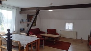 Un coin repas avec une table et des chaises en bois, un canapé rouge et une petite table surmontée d’un tapis rouge. Il y a une fenêtre avec des rideaux et un radiateur au mur.