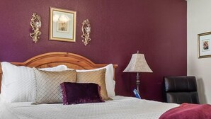 A bedroom with a wooden headboard, a bed with white and purple bedding, a nightstand with a lamp, and a ceiling fan.