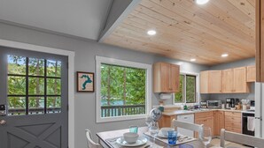 A modern kitchen with wooden cabinets, a dining area with a table and chairs, and large windows offering a view of trees.