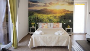Un dormitorio con una cama grande, un mural de paisaje al atardecer, mesitas de noche con lámparas y vistas al exterior a través de una ventana.