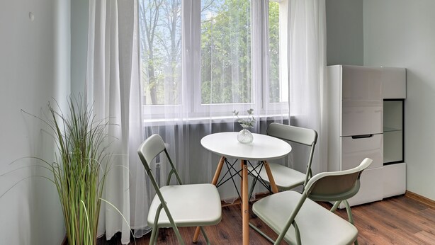 A small, bright room with a round table and four chairs, a white refrigerator, and a large window with sheer curtains. - RentPlanet - Apartament Piastowska (Gdansk)