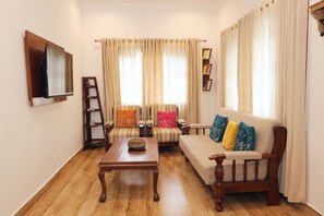 Ein Wohnzimmer mit Couchtisch aus Holz, zwei Sofas mit bunten Kissen, einem Bücherregal und einem Wand-Fernseher.