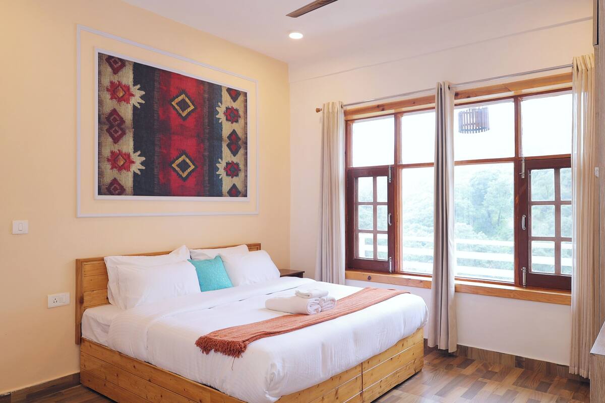 A neatly made bed with white linens and a folded towel, a wooden headboard, a large window with curtains, and a framed artwork on the wall.