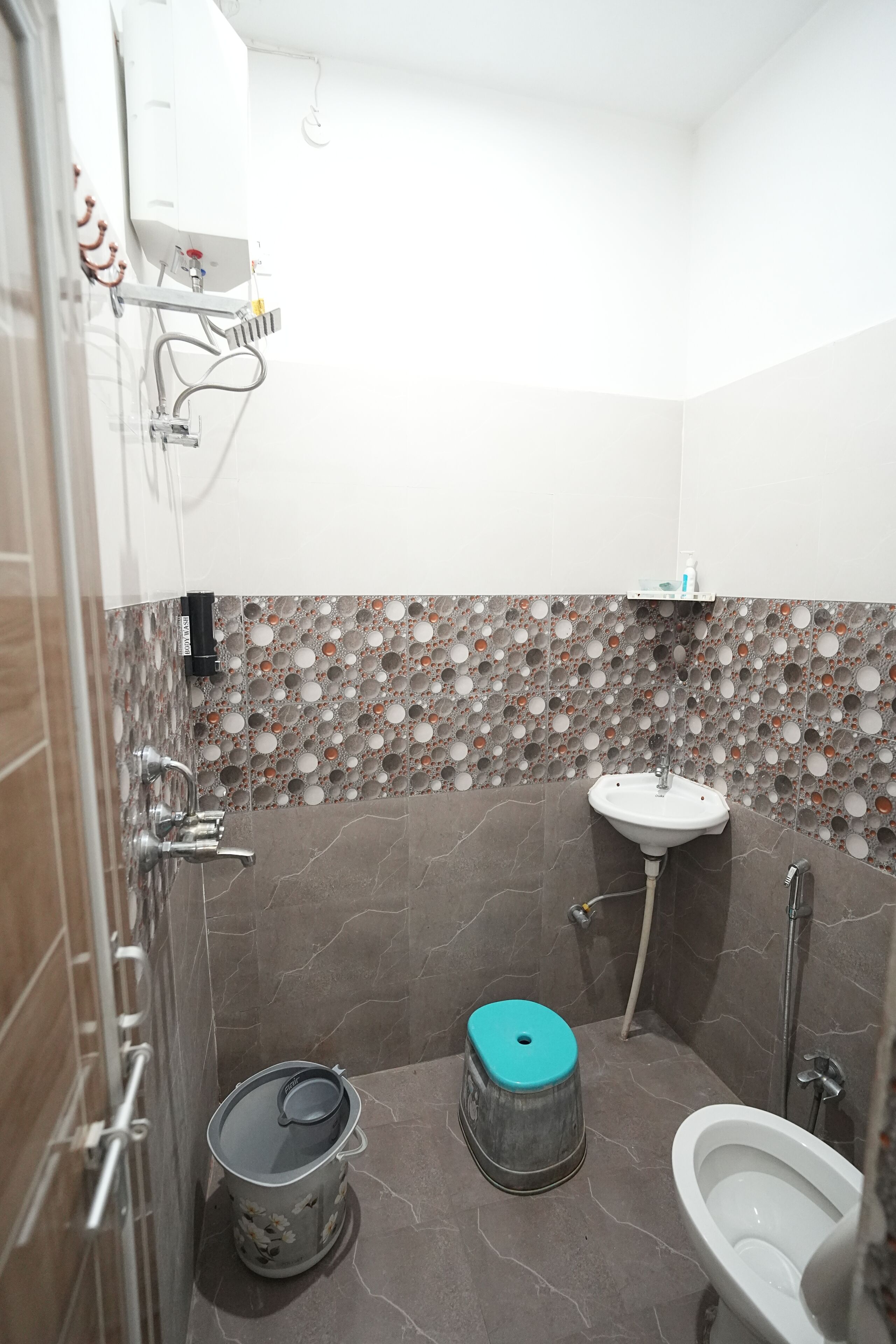 A bathroom with a toilet, sink, and shower. There is a trash can and a water heater on the wall.