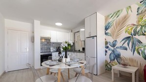 A modern kitchen with white cabinets, a dining table set for four, and a tropical leaf-patterned wall. - Andalucia 5 - One Bed (Costa Adeje)