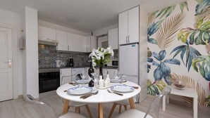 A modern kitchen with white cabinets, a dining table set for two, and a tropical leaf wallpaper. - Andalucia 5 - One Bed (Costa Adeje)