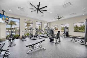 Une salle de sport équipée de divers appareils d’exercice, dotée de grandes fenêtres et d’un ventilateur de plafond.
