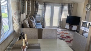 A modern living room with a sofa, television, and a dining area with a table set for two. - Seaside 2-bed Caravan Retreat - Sleeps 6 & Parking (Prestatyn)