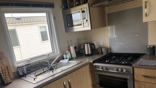A compact kitchen with a sink, stove, oven, microwave, and wooden cabinets. - Seaside 2-bed Caravan Retreat - Sleeps 6 & Parking (Prestatyn)