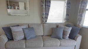 A living room with a grey sofa, patterned cushions, a bookshelf, and a window with curtains. - Seaside 2-bed Caravan Retreat - Sleeps 6 & Parking (Prestatyn)