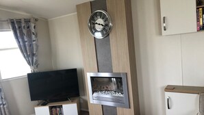A modern living room with a flat-screen TV, a fireplace, and a clock on the wall. - Seaside 2-bed Caravan Retreat - Sleeps 6 & Parking (Prestatyn)
