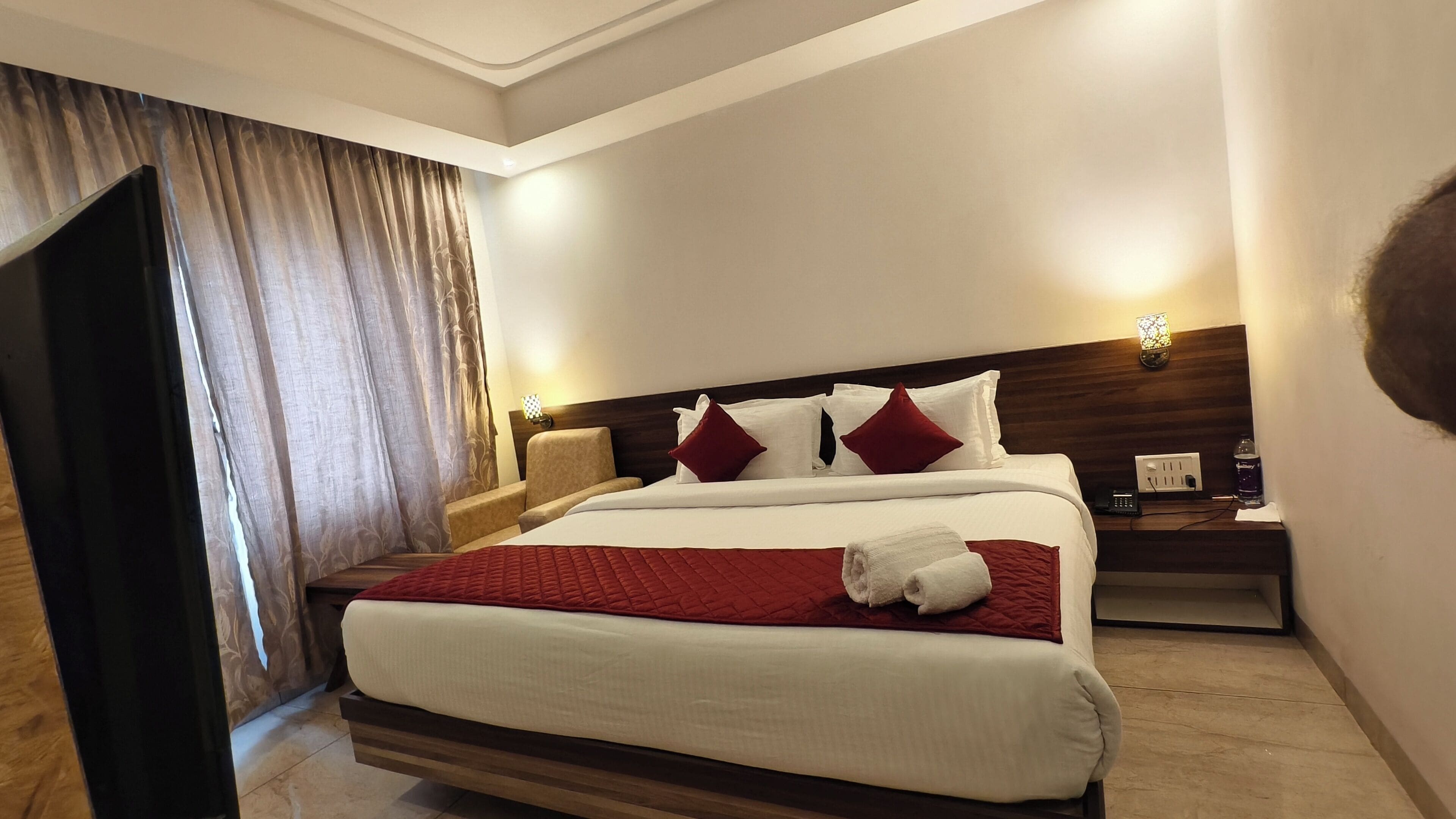 A hotel room with a large bed, a wooden headboard, a bedside table with a lamp, and a curtain.