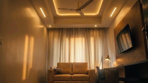 A room with a ceiling fan, recessed lighting, a sofa, a TV mounted on the wall, and a window with curtains. - Monarch Express, Sri City (Tada)