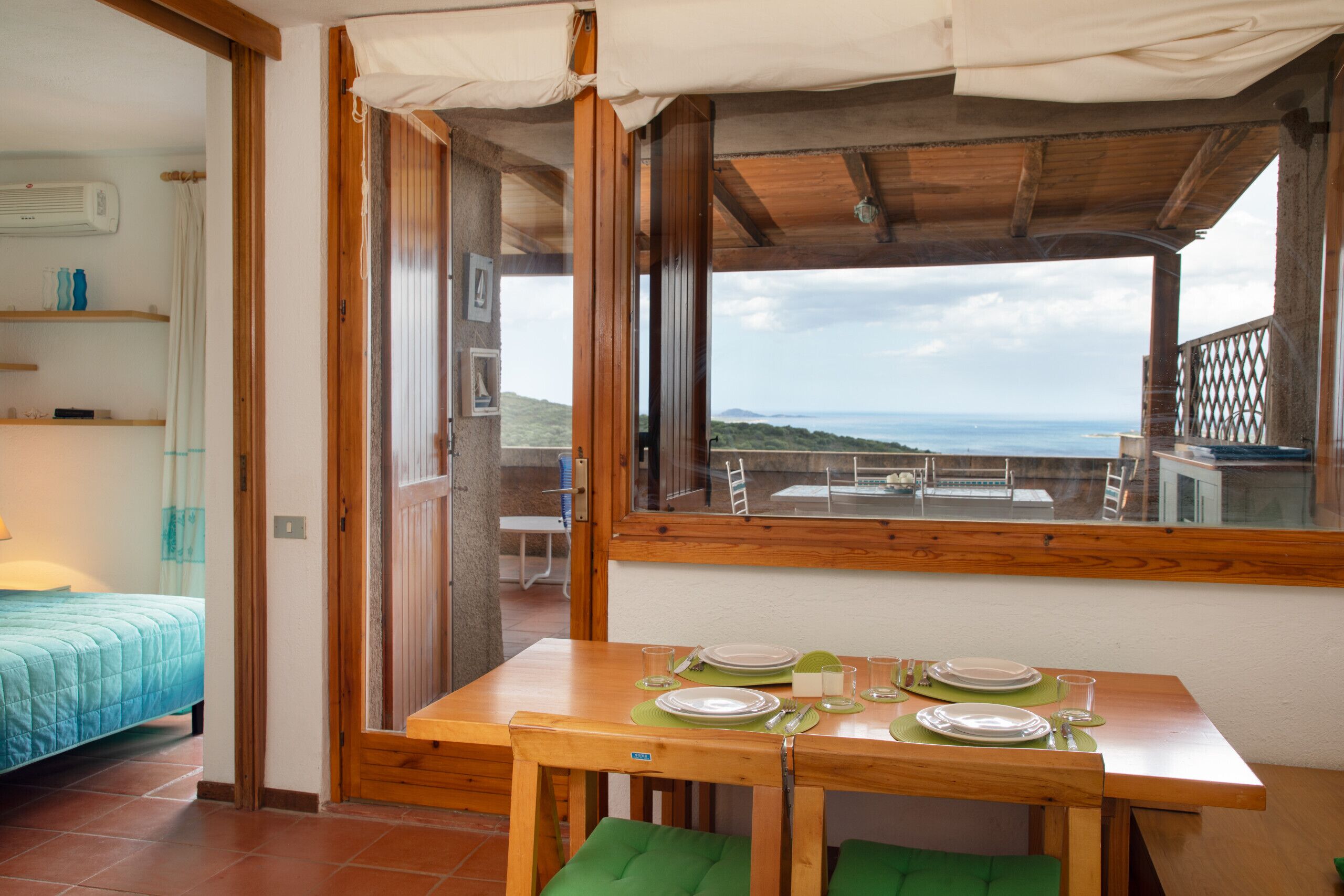 A room with a wooden table set for dining, a bed with a blue quilt, and a view of the sea through a large window.