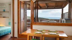 A room with a wooden table set for dining, a bed with a blue quilt, and a view of the sea through a large window. - Casa Sunrise by Wonderful Italy (Olbia)