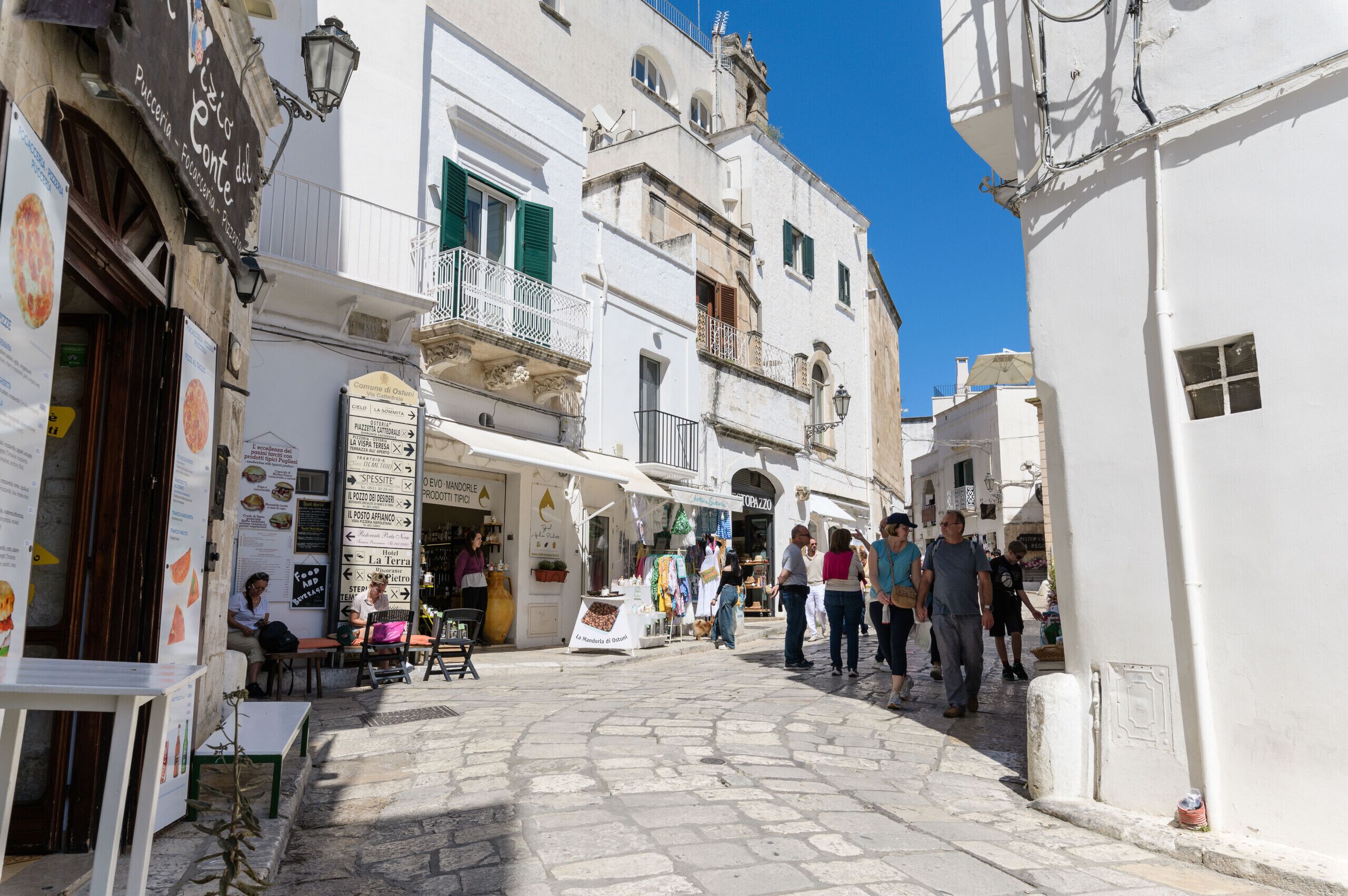 Une rue étroite bordée de bâtiments blancs, avec des terrasses et une variété de boutiques.