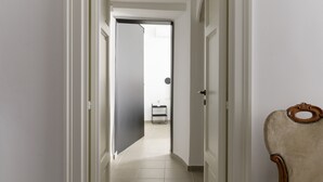 Een gang met een deur naar een kamer, waar een bed en een klein tafeltje staan.