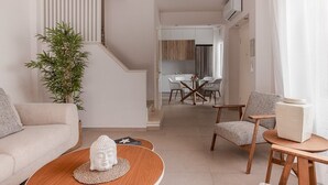 Un salon moderne avec un canapé, une table basse et des fauteuils, un escalier menant à un coin repas avec une table et des chaises, et une cuisine en arrière-plan.