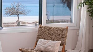 Une pièce avec vue sur la mer, grâce à une grande fenêtre, comprenant un fauteuil en osier avec un coussin, un lit recouvert d’une couverture à carreaux et une plante en pot.