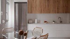 A modern kitchen with a stainless steel refrigerator, wooden cabinets, a glass dining table with wicker chairs, and a white countertop with a sink and a bowl of fruit. - Paradise Overlook - Beachfront Villa (Pefkohori)