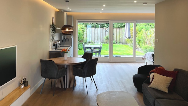 A modern living room with a dining area, a flat-screen TV, and a large sliding glass door leading to a backyard.