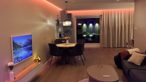 A modern living room with a flat-screen TV, a dining area with a table and chairs, a sofa, and a small round table.