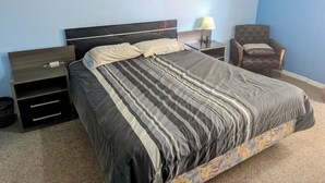 Ein Schlafzimmer mit einem Bett, einem Nachttisch, einem Sessel, einer Lampe und einem Bild an der Wand.