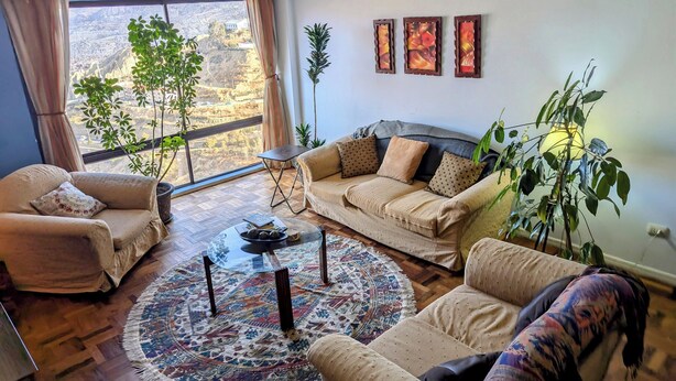 Ein Wohnzimmer mit einer Couch, einem Sessel und einem Couchtisch. An der Wand hängen drei Gemälde und ein großes Fenster mit Vorhängen.