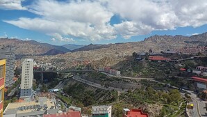 Eine Stadtansicht mit einem markanten Gebäude, einer Brücke und gebirgiger Landschaft im Hintergrund.
