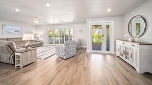 Un salon spacieux avec un canapé gris, un pouf à motifs et une table basse en bois. On y trouve une grande fenêtre avec des stores, un ventilateur de plafond et un meuble blanc avec un miroir.