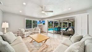 Un salon spacieux doté d’une grande fenêtre, d’un ventilateur de plafond et offrant une vue sur une piscine et un espace de détente extérieur.